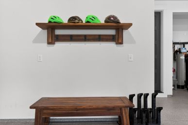 Garage entryway with rustic wooden shelf and hooks holding four bike helmets (two neon green), wooden bench below and black boot racks at the side.