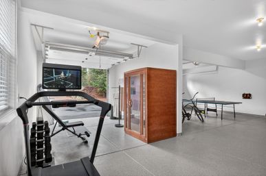 Bright residential garage converted into a home gym and game room with a treadmill and screen, adjustable dumbbells, weight bench, wooden sauna cabin, ping-pong table, bicycles, and an open garage door showing trees outside.