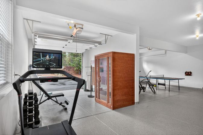 Bright residential garage converted into a home gym and game room with a treadmill and screen, adjustable dumbbells, weight bench, wooden sauna cabin, ping-pong table, bicycles, and an open garage door showing trees outside.