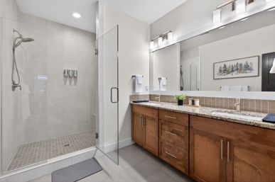Bright contemporary bathroom with glass walk-in shower and rain showerhead, neutral floor-to-ceiling tiles, granite double-sink vanity with wooden cabinets, large wall mirror, modern sconces and framed tree artwork.