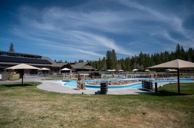 Sunny outdoor resort swimming pool and winding lazy river with stacked lounge chairs and umbrellas in front of a wooden mountain lodge surrounded by pine forest and blue sky.