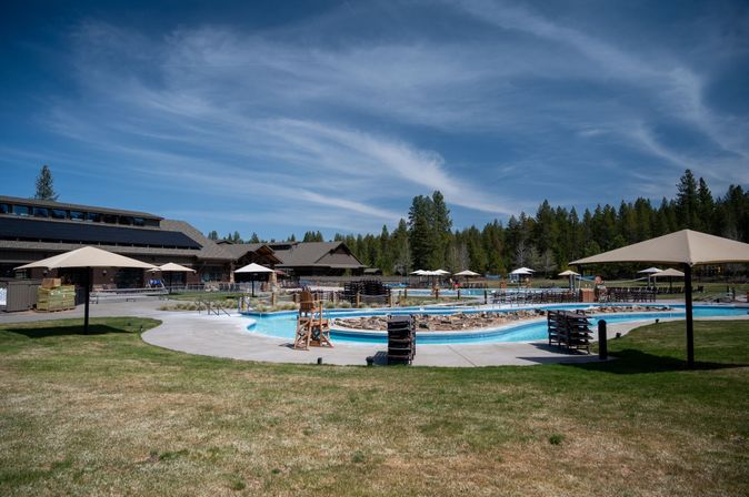 Sunny outdoor resort swimming pool and winding lazy river with stacked lounge chairs and umbrellas in front of a wooden mountain lodge surrounded by pine forest and blue sky.