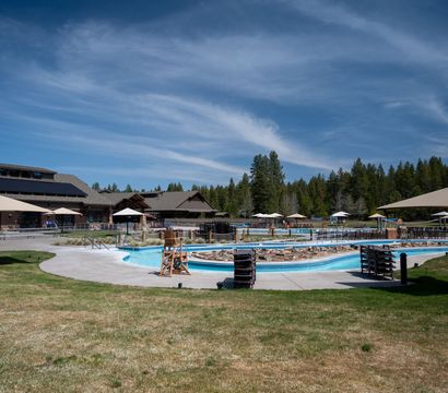 Sunny outdoor resort swimming pool and winding lazy river with stacked lounge chairs and umbrellas in front of a wooden mountain lodge surrounded by pine forest and blue sky.