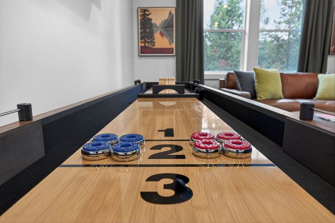 Close-up of a modern indoor shuffleboard table with blue and red pucks on the 1‑2‑3 scoring lanes, set in a bright living room with a brown leather sofa, green throw pillows and large windows overlooking trees.
