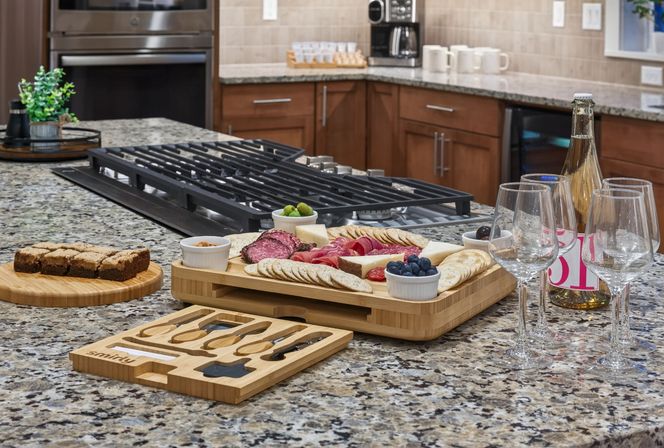 Modern kitchen island with granite countertop showcasing a wooden charcuterie board of cheeses, cured meats, crackers, olives and blueberries, three wine glasses and a bottle beside a built-in gas cooktop — ready for entertaining.