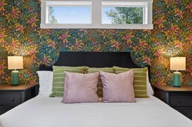 Cozy bedroom with black headboard, playful floral wallpaper, green striped pillows, pink velvet cushions, matching green bedside lamps and a narrow high window with a tree view.
