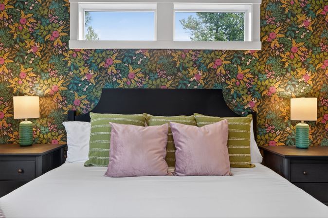 Cozy bedroom with black headboard, playful floral wallpaper, green striped pillows, pink velvet cushions, matching green bedside lamps and a narrow high window with a tree view.
