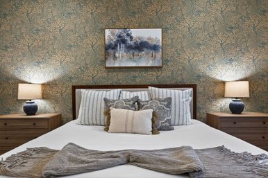 Cozy bedroom interior with made bed, striped and patterned pillows and a knitted throw, wooden nightstands with lamps, blue-gold botanical wallpaper and framed forest artwork