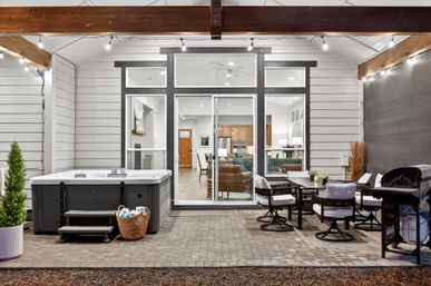 Covered backyard patio with hot tub and wicker towel basket, outdoor dining set and grill under string lights, sliding glass doors revealing a modern living room and kitchen
