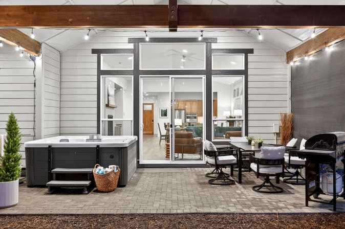 Covered backyard patio with hot tub and wicker towel basket, outdoor dining set and grill under string lights, sliding glass doors revealing a modern living room and kitchen