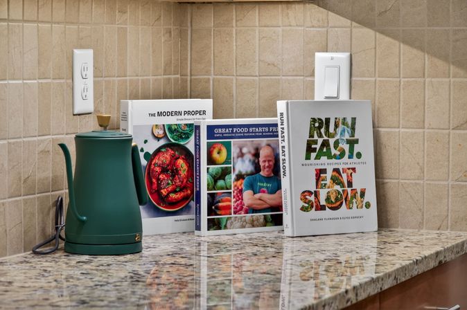 Cozy kitchen countertop vignette: matte green electric kettle on speckled granite with beige tile backsplash, three upright cookbooks with colorful food photos and a bold "RUN FAST. EAT SLOW." cover.