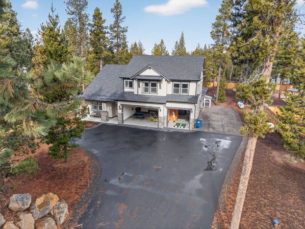 Aerial view of a two-story gray house nestled among tall pine trees, large asphalt driveway leading to an open two-car garage with bikes and a game table, forested residential property.