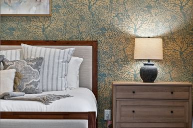 Cozy bedroom scene with a wooden bed frame, striped and textured throw pillows, gray fringed blanket, teal-and-gold floral wallpaper, and a wooden nightstand topped by a glowing ceramic table lamp.