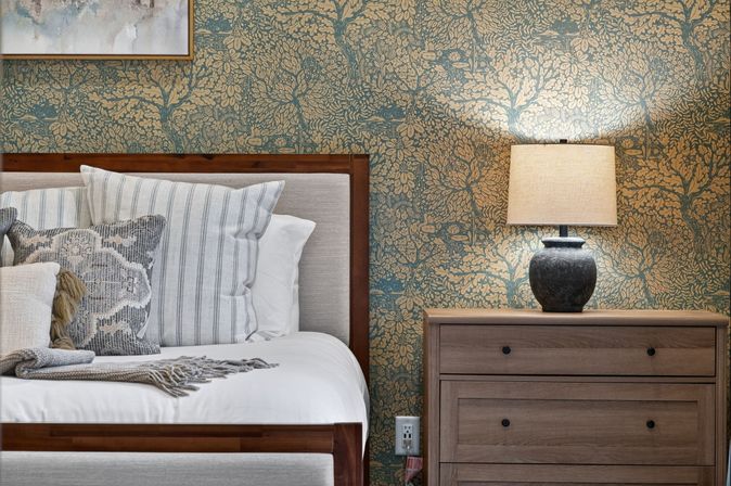 Cozy bedroom scene with a wooden bed frame, striped and textured throw pillows, gray fringed blanket, teal-and-gold floral wallpaper, and a wooden nightstand topped by a glowing ceramic table lamp.