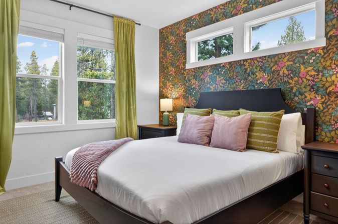 Cozy forest-view bedroom with dark wood bed, crisp white linens, pastel pink throw and pillows, green striped accent pillows, floral wallpaper accent wall, green velvet curtains, bedside tables with lamp, and large windows showing pine trees outside.
