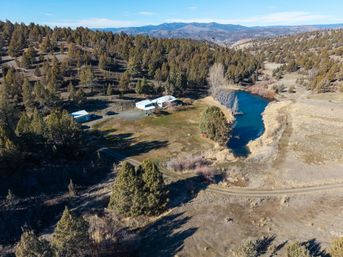 Aerial shot of a cozy rural ranch beside a vivid blue pond, outbuildings and a winding dirt road amid juniper-covered rolling hills and distant mountains