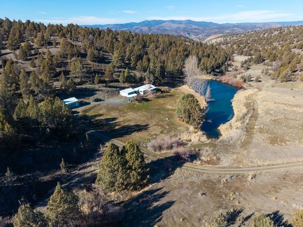 Aerial shot of a cozy rural ranch beside a vivid blue pond, outbuildings and a winding dirt road amid juniper-covered rolling hills and distant mountains