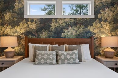 Cozy rustic bedroom with wooden headboard, dark botanical wallpaper, layered neutral pillows and textured lumbar cushions, matching bedside tables and warm lamps beneath a small high window showing pine trees.