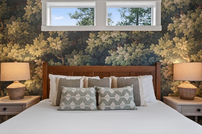 Cozy rustic bedroom with wooden headboard, dark botanical wallpaper, layered neutral pillows and textured lumbar cushions, matching bedside tables and warm lamps beneath a small high window showing pine trees.