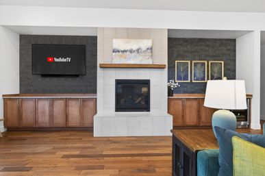 Modern open living room with central tiled fireplace and wooden mantel, wall-mounted flat-screen TV displaying YouTube TV logo, dark textured accent walls with built-in wood cabinets, hardwood floors, teal sofa and table lamp in foreground.