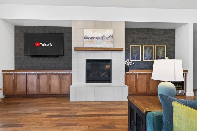 Modern open living room with central tiled fireplace and wooden mantel, wall-mounted flat-screen TV displaying YouTube TV logo, dark textured accent walls with built-in wood cabinets, hardwood floors, teal sofa and table lamp in foreground.
