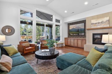 Bright modern living room with teal corduroy sectional, two tan leather armchairs, round wooden coffee table with potted greenery, floor-to-ceiling windows overlooking a backyard patio, built-in wood media cabinets, wall-mounted TV above a fireplace, and warm hardwood floors.