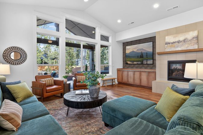 Bright modern living room with teal corduroy sectional, two tan leather armchairs, round wooden coffee table with potted greenery, floor-to-ceiling windows overlooking a backyard patio, built-in wood media cabinets, wall-mounted TV above a fireplace, and warm hardwood floors.