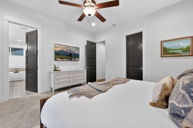 Contemporary master bedroom with white walls, ceiling fan, king bed with textured pillows and throw, white dresser and wall-mounted TV, open door to tiled ensuite bathroom