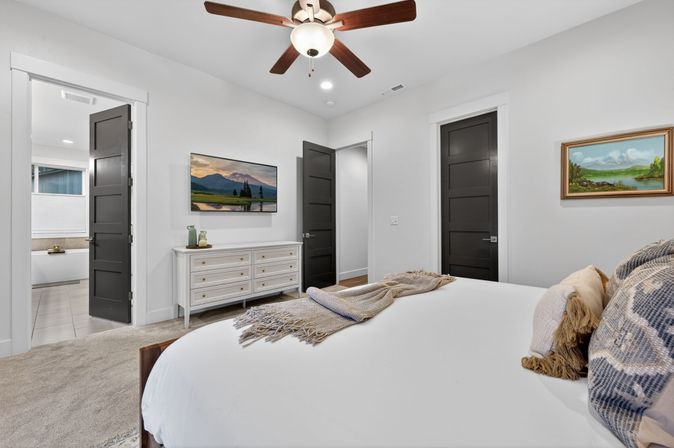 Contemporary master bedroom with white walls, ceiling fan, king bed with textured pillows and throw, white dresser and wall-mounted TV, open door to tiled ensuite bathroom