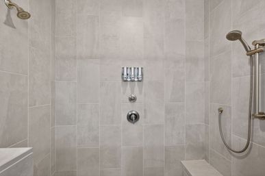 Spa-ready walk-in shower with sleek neutral gray rectangular tiles, built-in bench, overhead and handheld showerheads, and three wall-mounted soap dispensers.