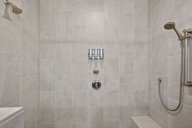 Spa-ready walk-in shower with sleek neutral gray rectangular tiles, built-in bench, overhead and handheld showerheads, and three wall-mounted soap dispensers.