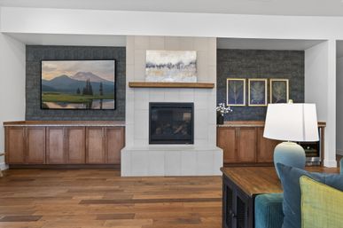 Stylish living room with central tiled fireplace and wooden mantel, wall-mounted TV showing a mountain landscape, flanking built-in wood cabinets on a dark textured accent wall, hardwood floors, lamp and teal sofa.