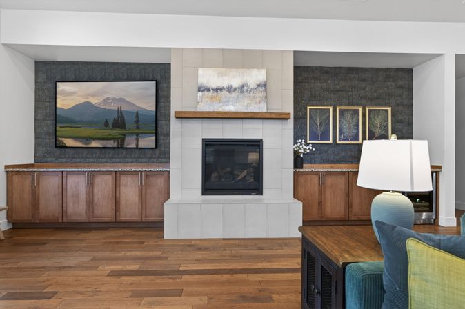 Stylish living room with central tiled fireplace and wooden mantel, wall-mounted TV showing a mountain landscape, flanking built-in wood cabinets on a dark textured accent wall, hardwood floors, lamp and teal sofa.