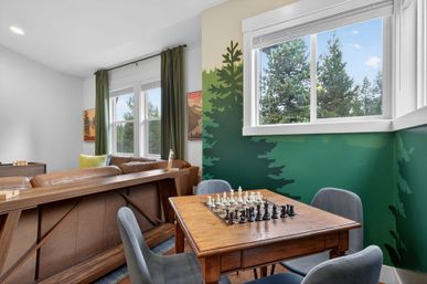 Cozy game corner in a modern living room — wooden chess table with pieces and gray chairs, brown leather sofa, green forest mural on the wall, and a large window framing evergreen trees and blue sky.