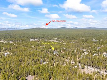 Aerial view of a vast pine forest with scattered homes and dirt roads; a yellow arrow marks a cabin cluster and a red arrow points to snow‑capped Mt. Bachelor on the distant horizon under a bright blue sky with fluffy clouds.