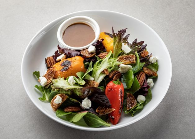 Grilled peach and roasted red pepper salad on mixed baby greens with dried figs, candied pecans, crumbled goat cheese and balsamic vinaigrette in a white bowl