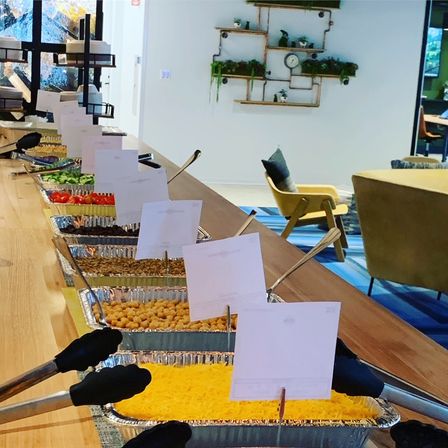 Salad-bar style toppings in foil pans with tongs and blank labels on a long wooden counter in a modern coworking lounge with chairs and wall planters.