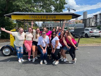 Group of smiling women in bachelorette tees, sashes and heart sunglasses posing in front of a yellow open-air party trolley on a sunny urban street and parking lot