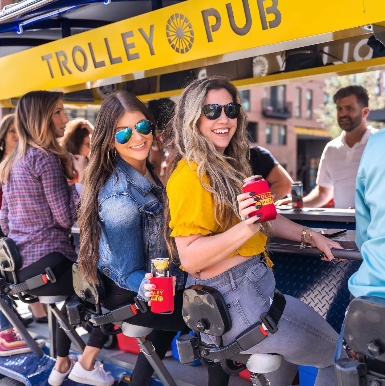 Two smiling women in sunglasses riding a yellow trolley bar in downtown, holding canned drinks in red koozies during an outdoor pub crawl.
