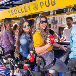 Two smiling women in sunglasses riding a yellow trolley bar in downtown, holding canned drinks in red koozies during an outdoor pub crawl.