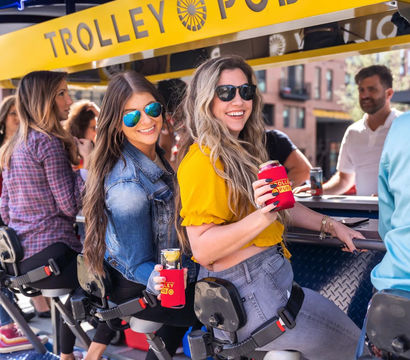 Two smiling women in sunglasses riding a yellow trolley bar in downtown, holding canned drinks in red koozies during an outdoor pub crawl.