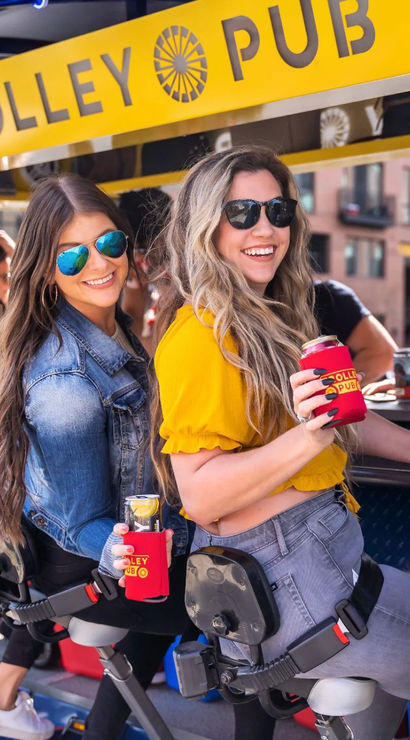 Two smiling women in sunglasses riding a yellow trolley bar in downtown, holding canned drinks in red koozies during an outdoor pub crawl.