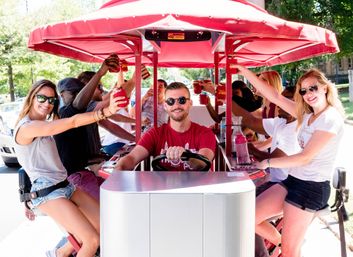 Group of friends smiling on a red pedal pub (party bike) led by a driver, raising drinks on a sunny tree-lined street during a daytime city bike tour