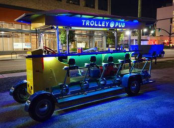 Brightly lit pedal-powered party trolley parked on a downtown street at night, long bar with bike seats and stools under blue-green LED lighting creating a festive urban vibe.