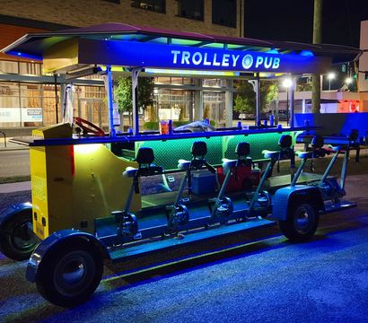 Brightly lit pedal-powered party trolley parked on a downtown street at night, long bar with bike seats and stools under blue-green LED lighting creating a festive urban vibe.