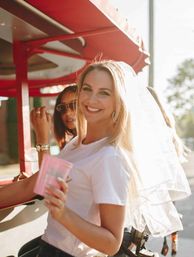 Smiling bride-to-be in a veil and white tee holding a pink cup during a sunny outdoor bachelorette party with friends