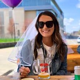 Smiling woman wearing sunglasses and a bachelorette veil holding a beer mug at a sunny outdoor patio with a purple balloon and city buildings in the background.