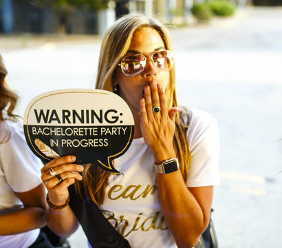 Bride-to-be in sunglasses holding a speech-bubble sign reading 'WARNING: Bachelorette party in progress' with friends smiling at an outdoor celebration