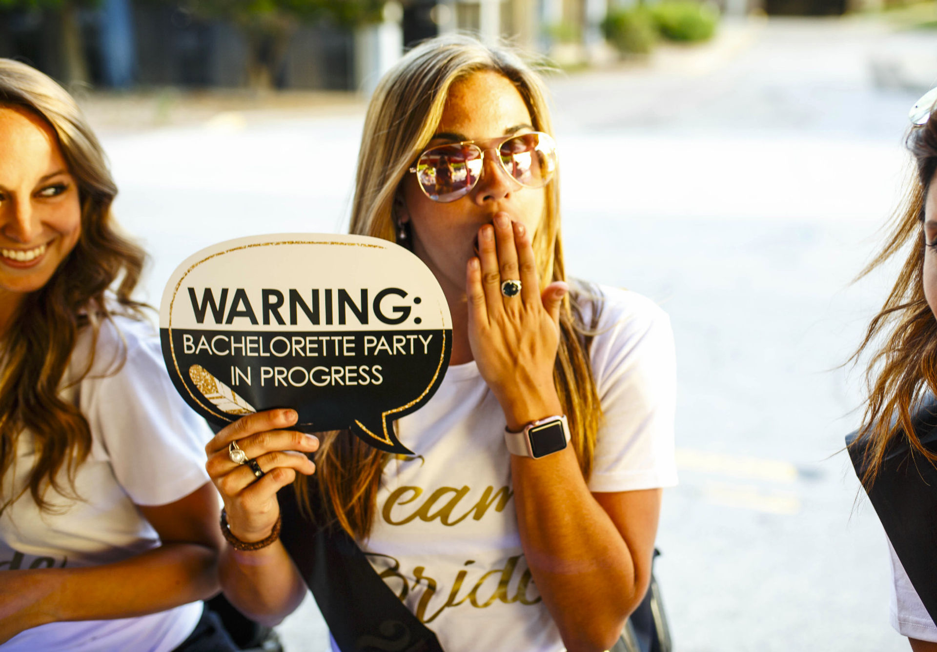 Bride-to-be in sunglasses holding a speech-bubble sign reading 'WARNING: Bachelorette party in progress' with friends smiling at an outdoor celebration