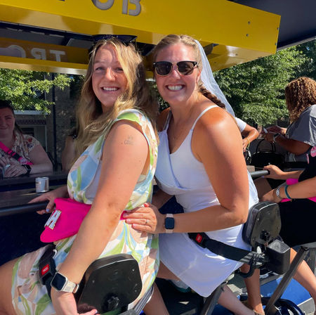 Two smiling women seated on an outdoor party bike under a yellow canopy, one wearing a veil and sunglasses, enjoying a sunny bachelorette celebration with friends on a city street.